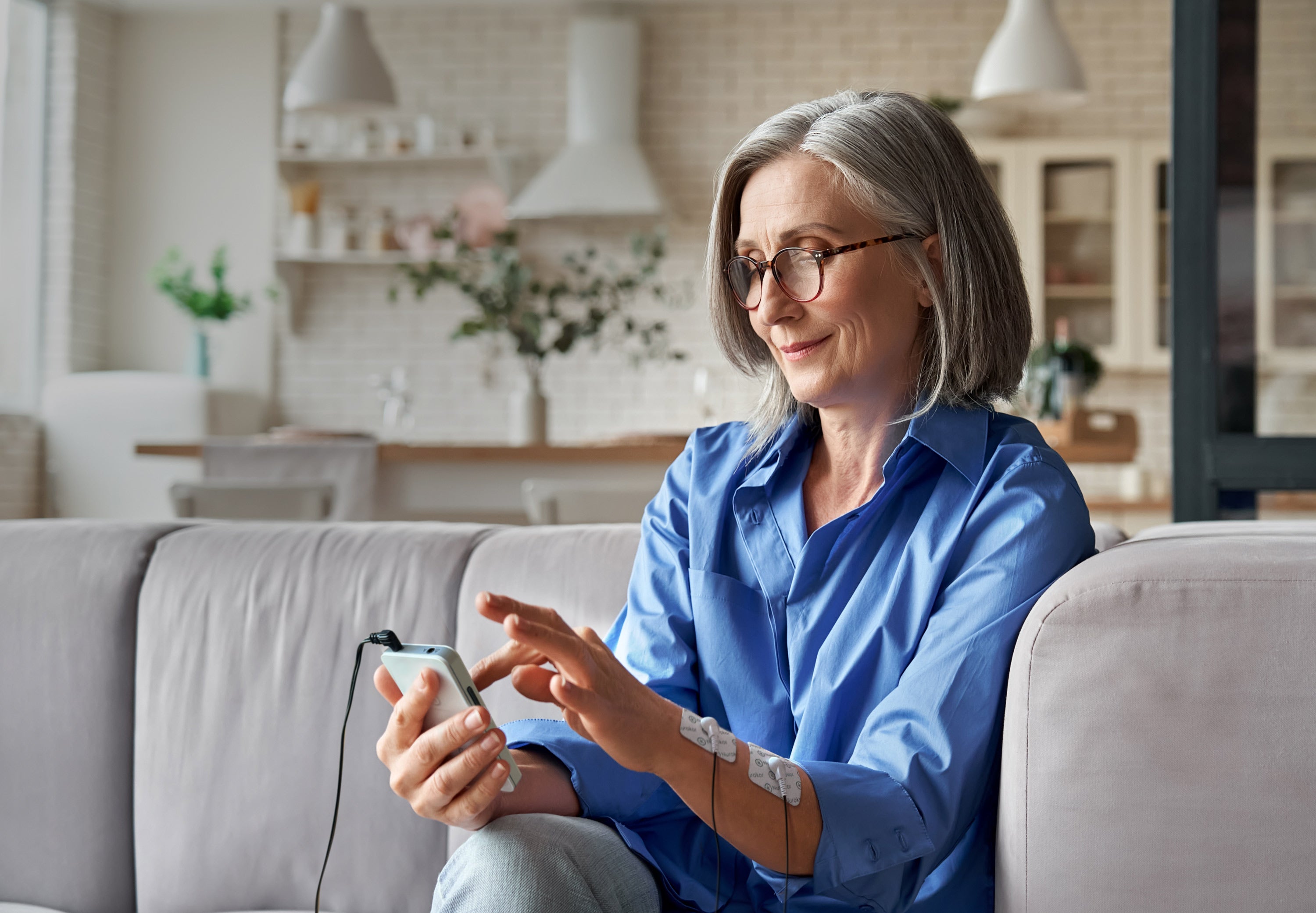 Middle aged lady on couch using the mitouch device with electrode pads on her forearm, smiling, managing pain, pain management