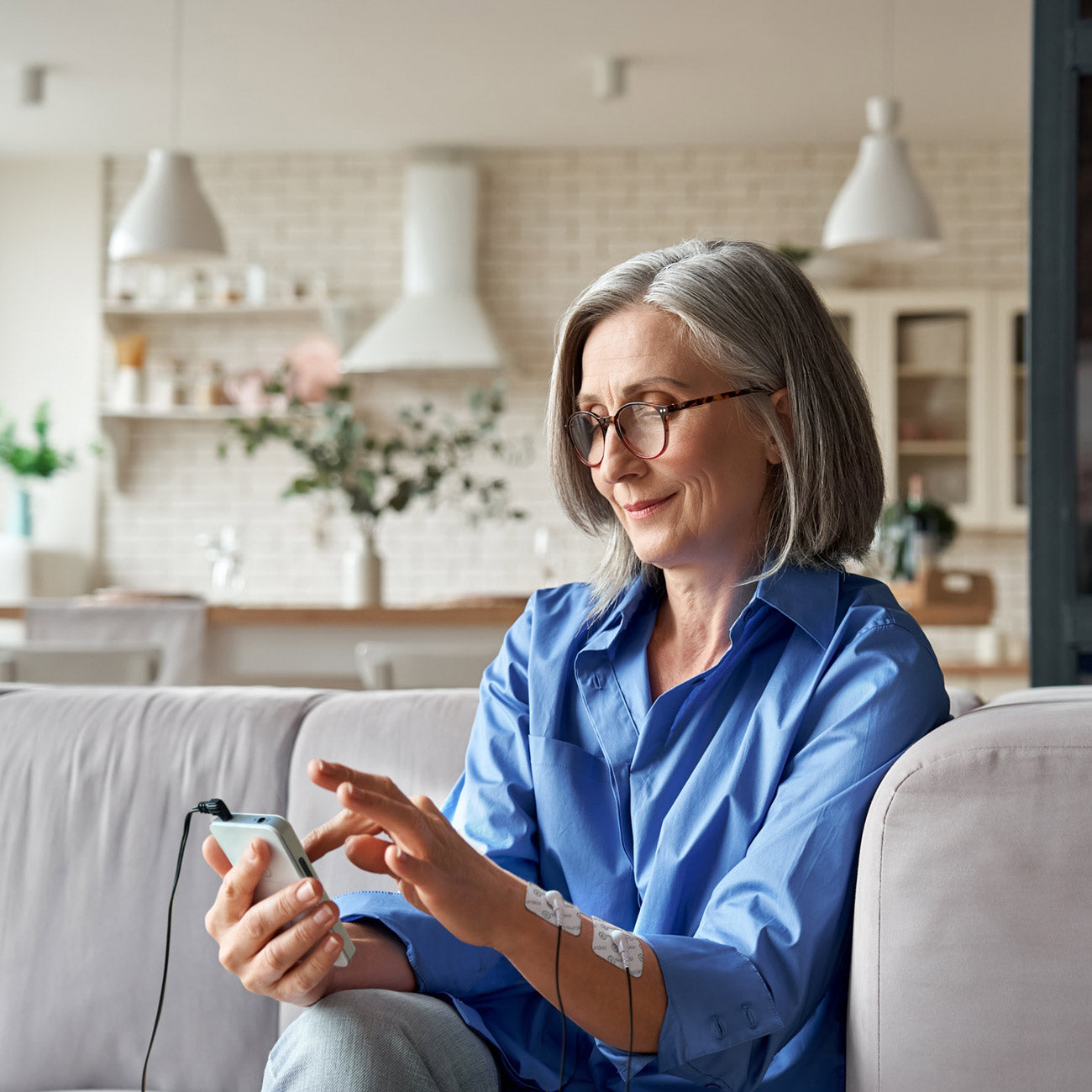 Middle aged lady on couch using the mitouch device with electrode pads on her forearm, smiling, managing pain, pain management