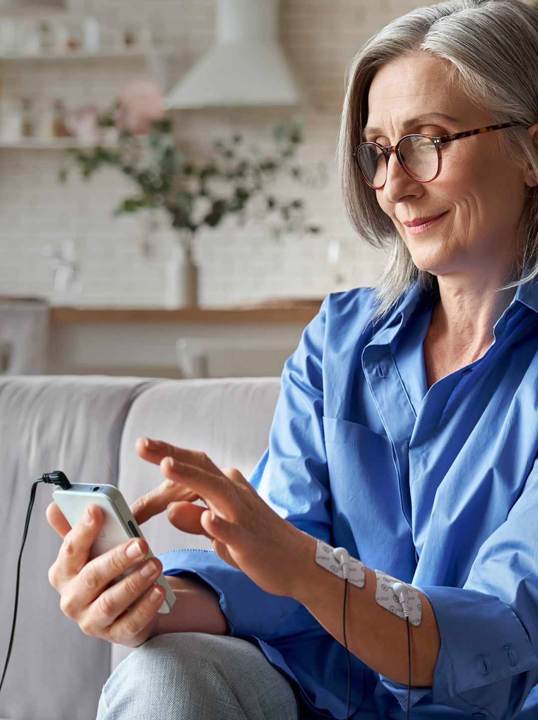 Middle aged lady on couch using the mitouch device with electrode pads on her forearm, smiling, managing pain, pain management